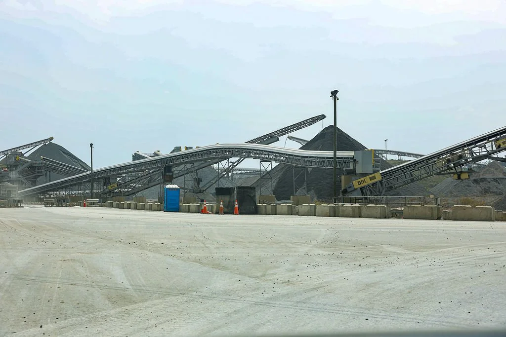 Industrial site with conveyor belts transporting black gravel or coal, large piles of material, crates, and a portable toilet, under a cloudy sky.