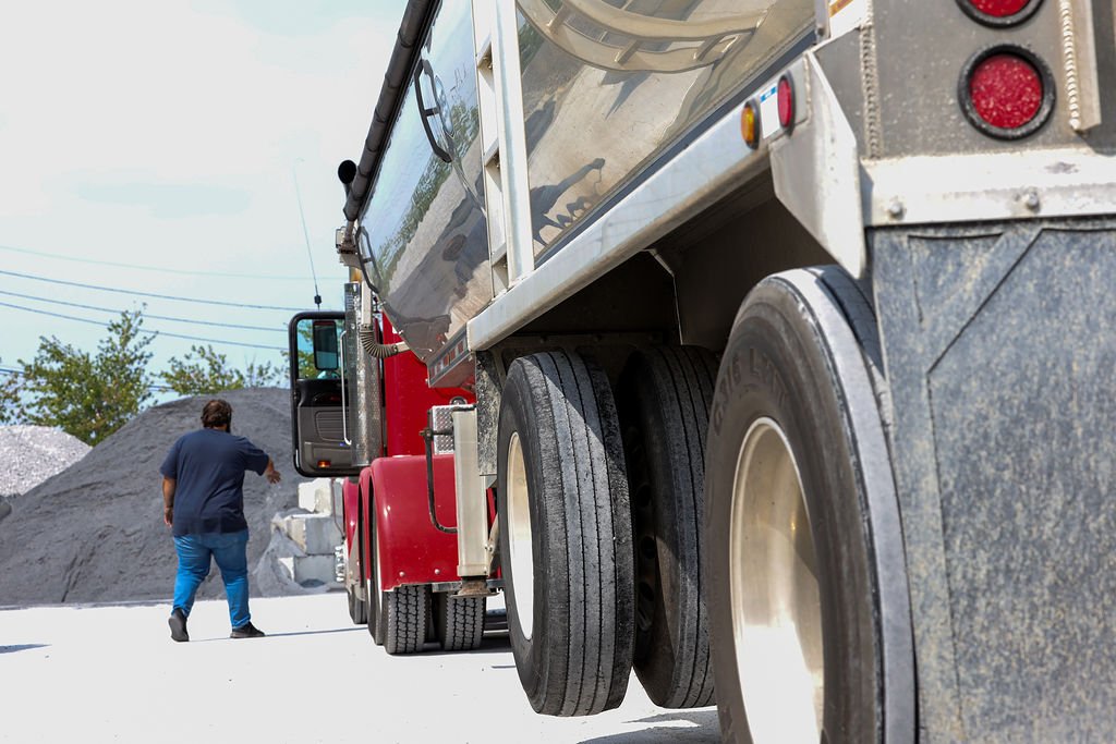 Creekstone red and silver multi-axle dump truck parked on a concrete surface as a worker approaches near a gravel pile, with trees and power lines in the background, highlighting heavy hauling and construction site operations.