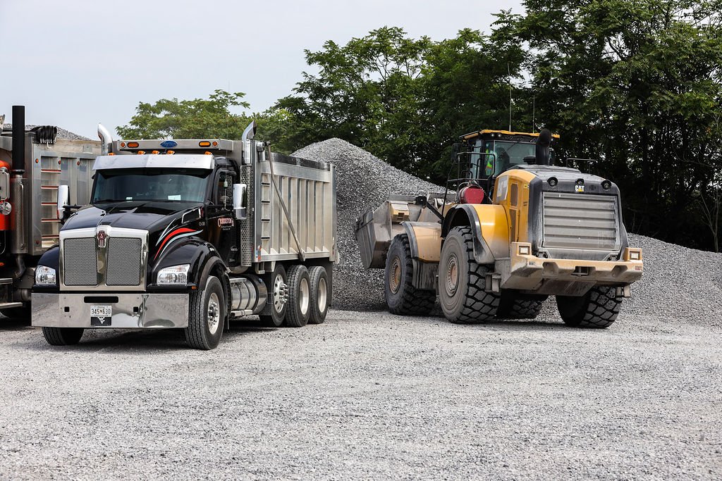 Creekstone black semi-truck parked beside a yellow bulldozer on a gravel lot with trees in the background, emphasizing heavy equipment, trucking, and construction site operations.