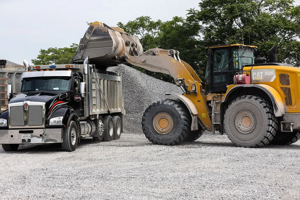 Creekstone yellow Caterpillar front loader moving gravel into a black and gray dump truck at a construction site with trees in the background, highlighting heavy equipment operation and material hauling services.