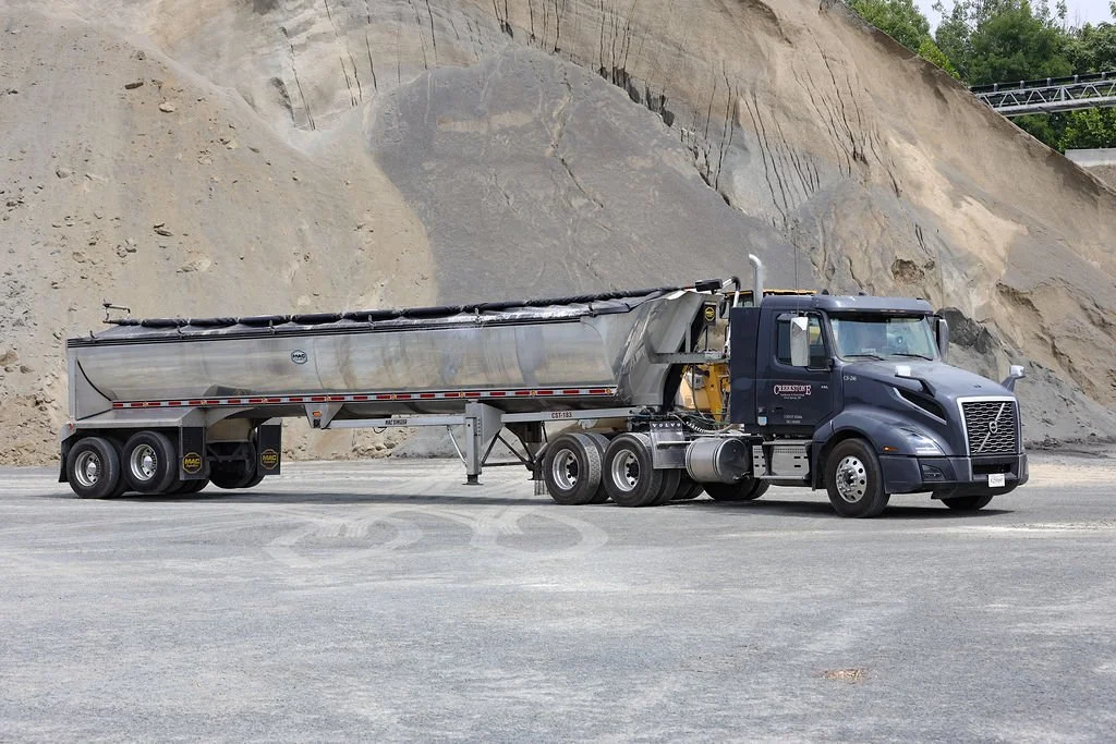 Creekstone black semi-truck with a silver trailer parked on a gravel lot in front of a large mountain of dirt and gravel, highlighting heavy hauling and material transport services.