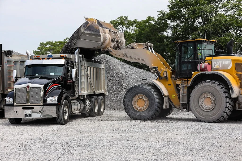 Creekstone red semi-truck with a chrome grille and bumper parked on a construction site being loaded with gravel from a loader.