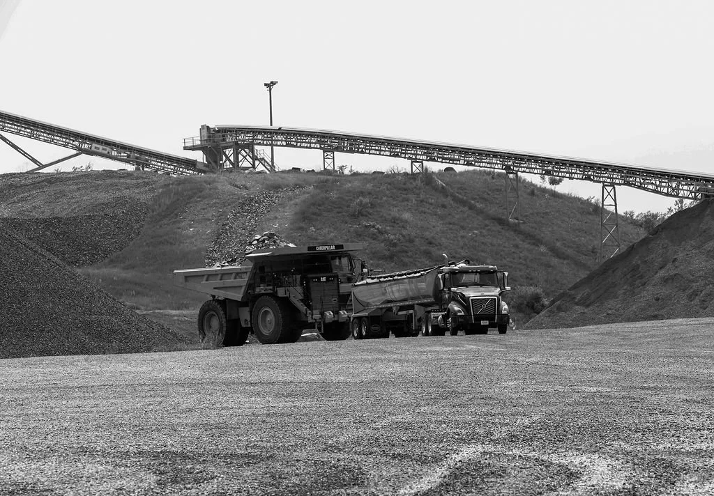 Creekstone industrial site with trucks, large gravel piles, an industrial conveyor belt, and a hillside in the background, highlighting aggregate production and heavy hauling operations.