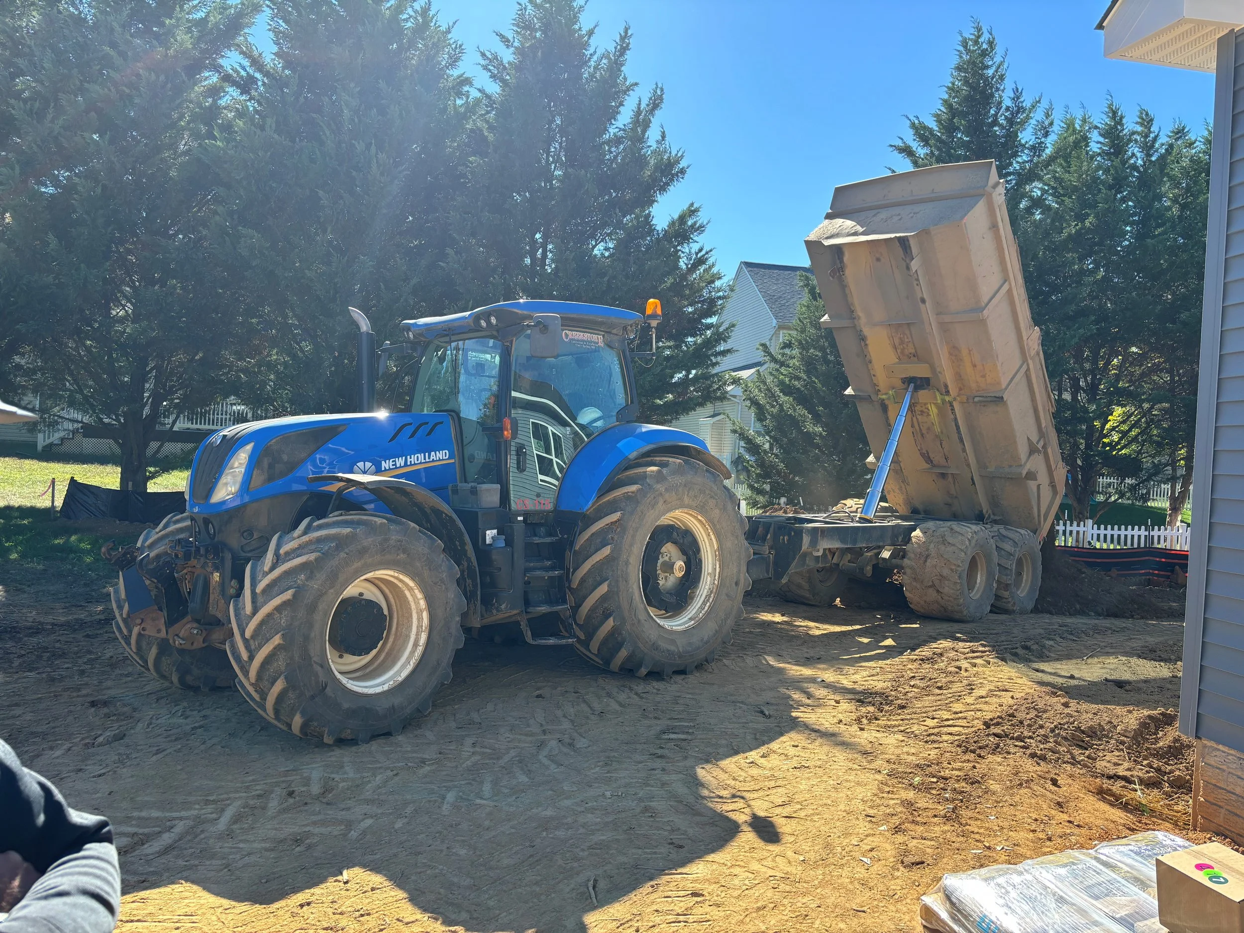 A blue New Holland tractor with large tires is on a dirt construction site, with its dump bed raised, dumping dirt onto the ground. There are trees and houses in the background.