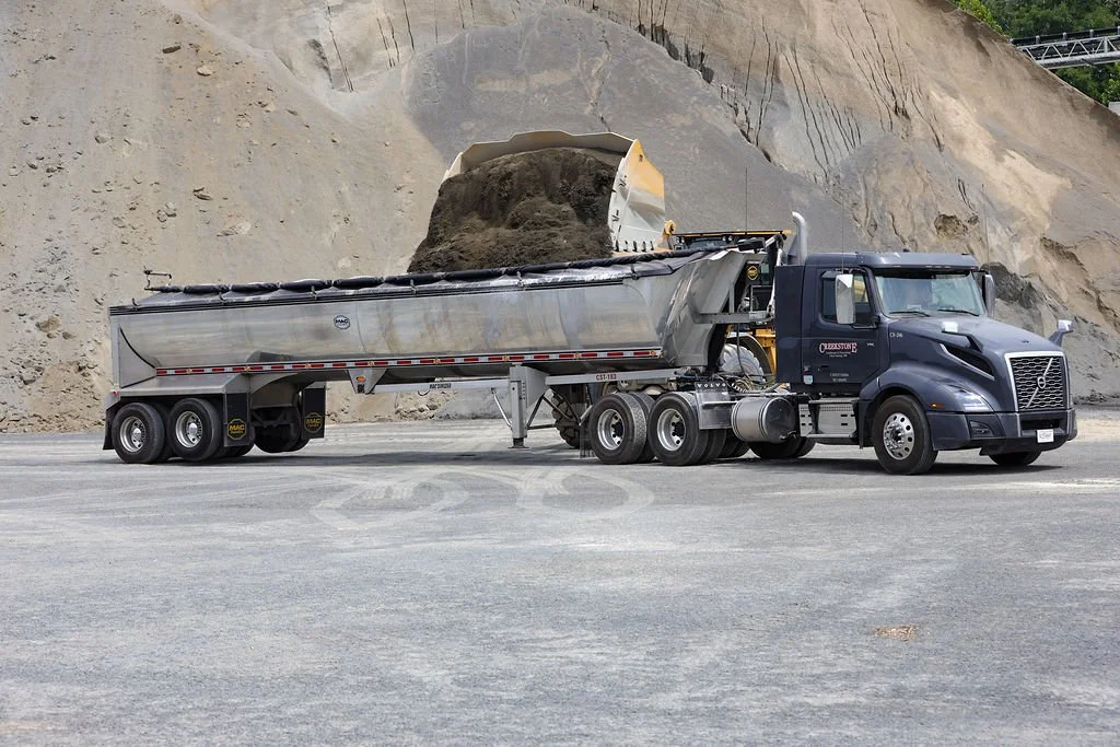 Creekstone black semi-truck with a front scoop loading dirt into its trailer at a construction site with large dirt piles in the background, emphasizing heavy hauling and site-work services.