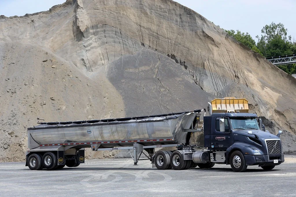 Creekstone black and silver semi-truck with a tanker trailer parked on a road in front of a large dirt and rock embankment, highlighting heavy-duty trucking and material transport services.