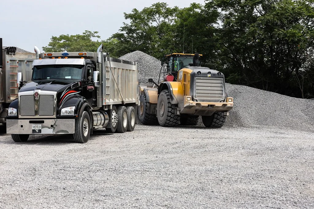 A black dump truck and a yellow front loader on a gravel surface with piles of gravel and trees in the background.