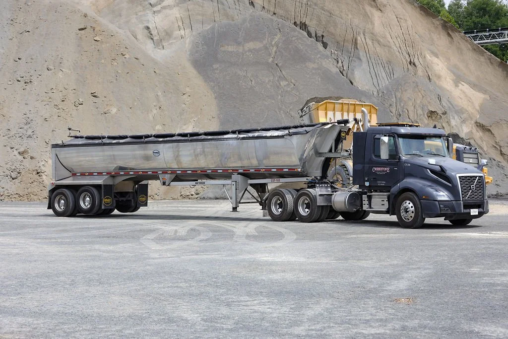 Creekstone black Volvo semi-truck with a silver tanker trailer parked on a flat gravel surface with a rocky hillside in the background, highlighting heavy-duty trucking and material transport services.