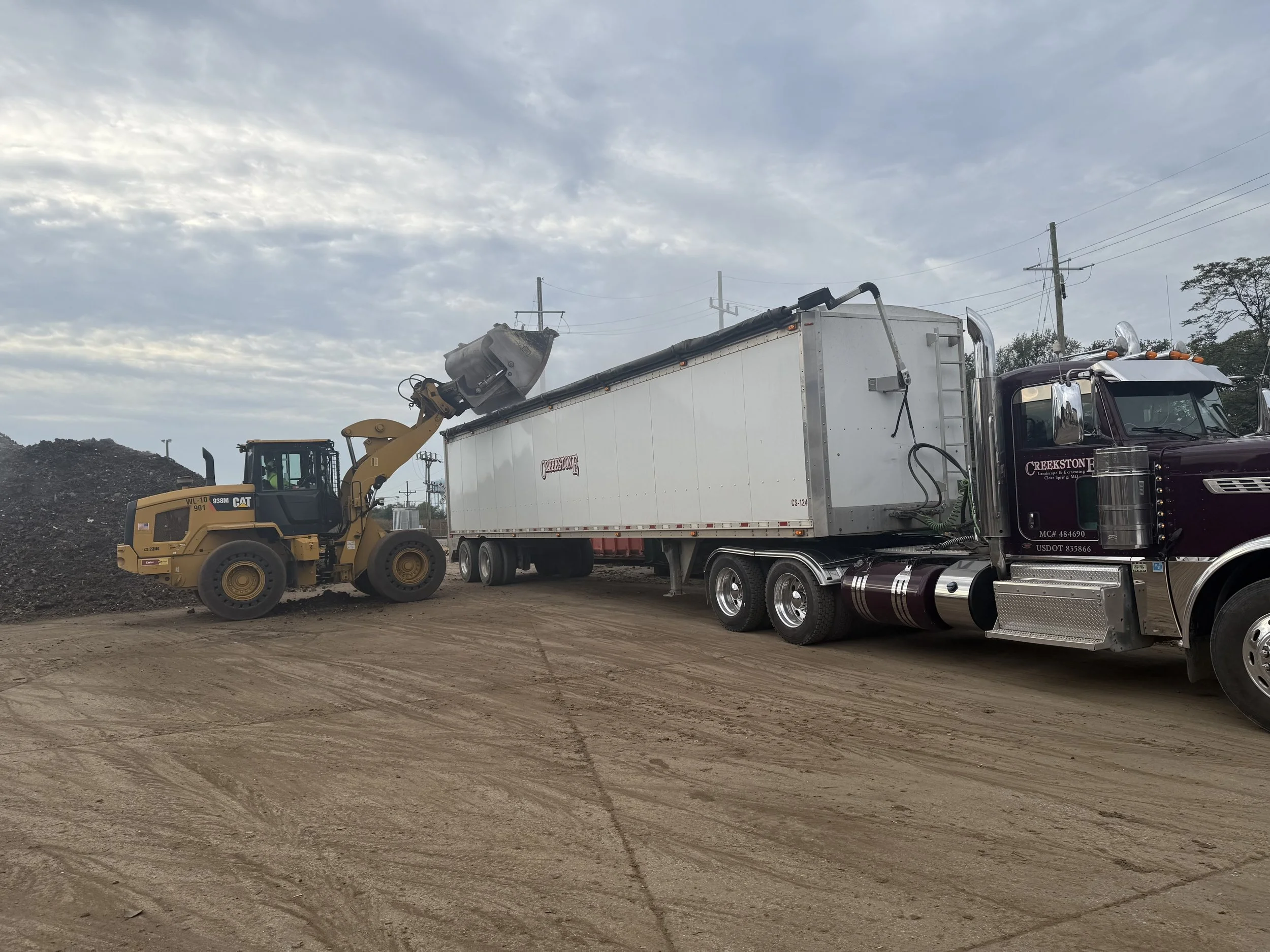 Creekstone bulldozer loading soil into a large semi-truck with a white cargo container on a dirt construction site, highlighting excavation, material handling, and heavy hauling services.