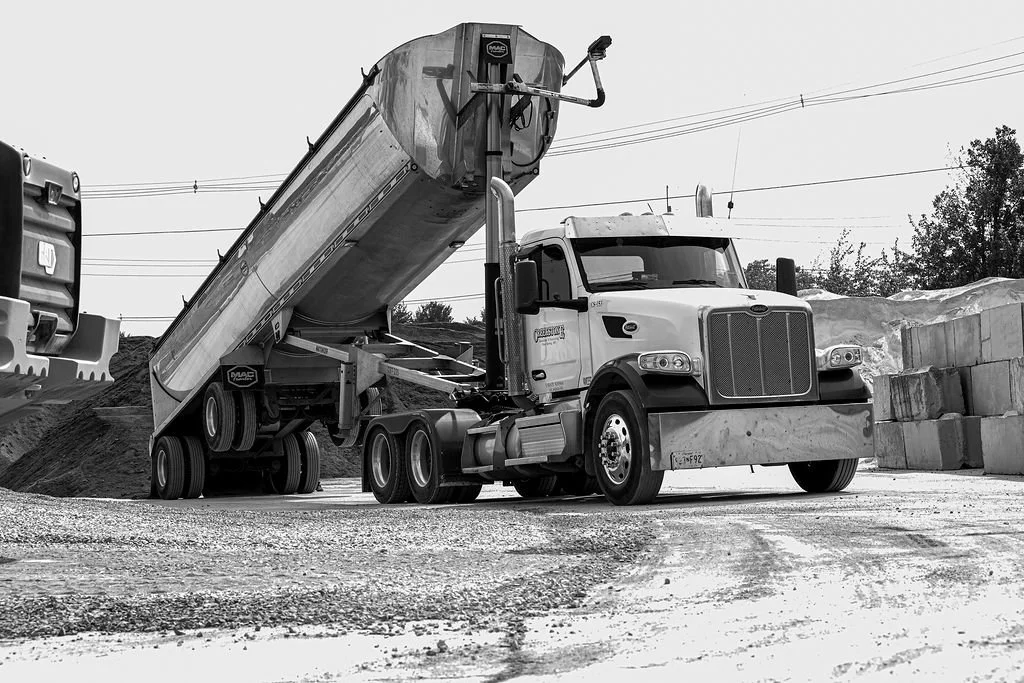 Creekstone semi-truck with a large hopper trailer tilted upward at a construction site, with concrete blocks on the right and another truck in the background, highlighting heavy hauling and material handling services.