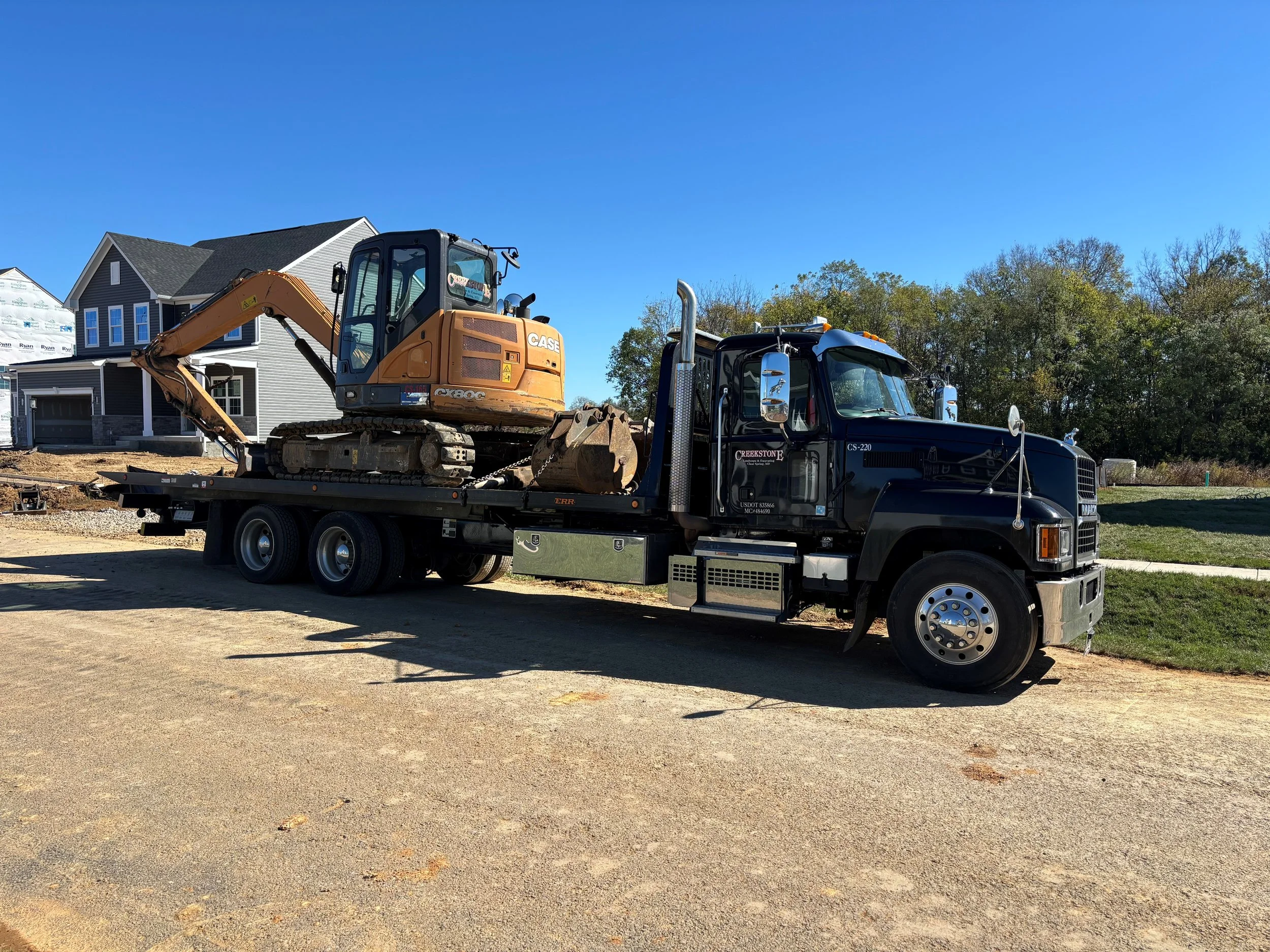A black flatbed truck carrying a small orange CASE excavator on a construction site with residential houses in the background and a clear blue sky.