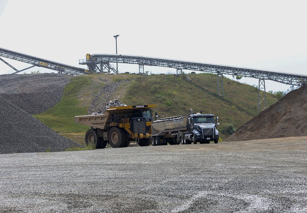 Creekstone industrial gravel quarry with large dump trucks and a conveyor system on a gravel surface with hilly terrain in the background, highlighting aggregate production and heavy hauling operations.