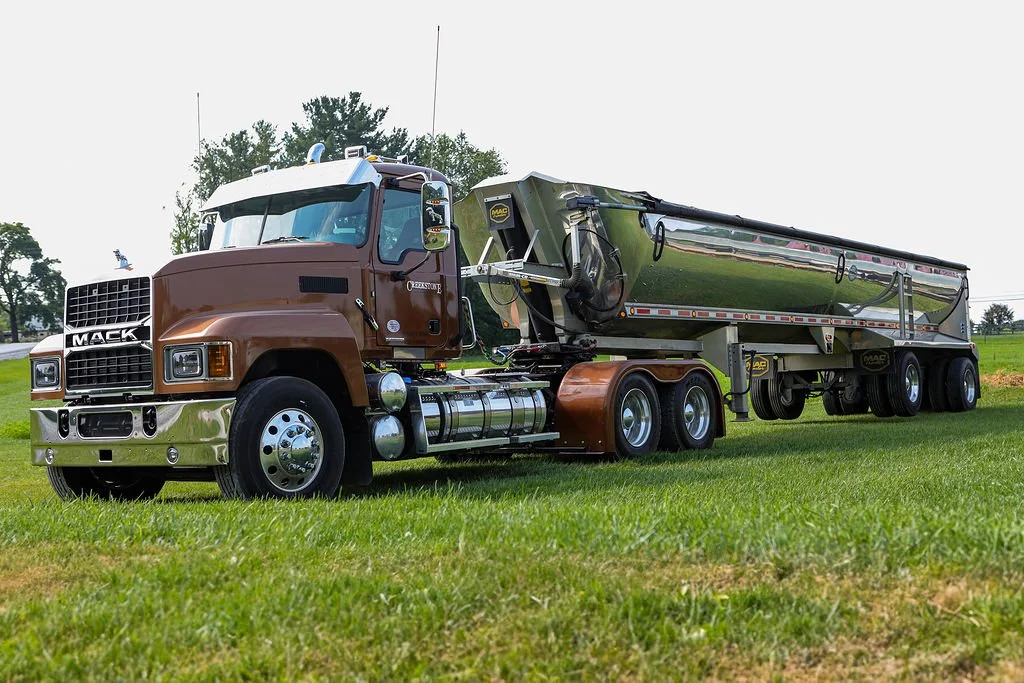 Creekstone Mack semi-truck with a brown cab and shiny silver trailer parked on a grassy field, highlighting heavy-duty trucking and hauling services.