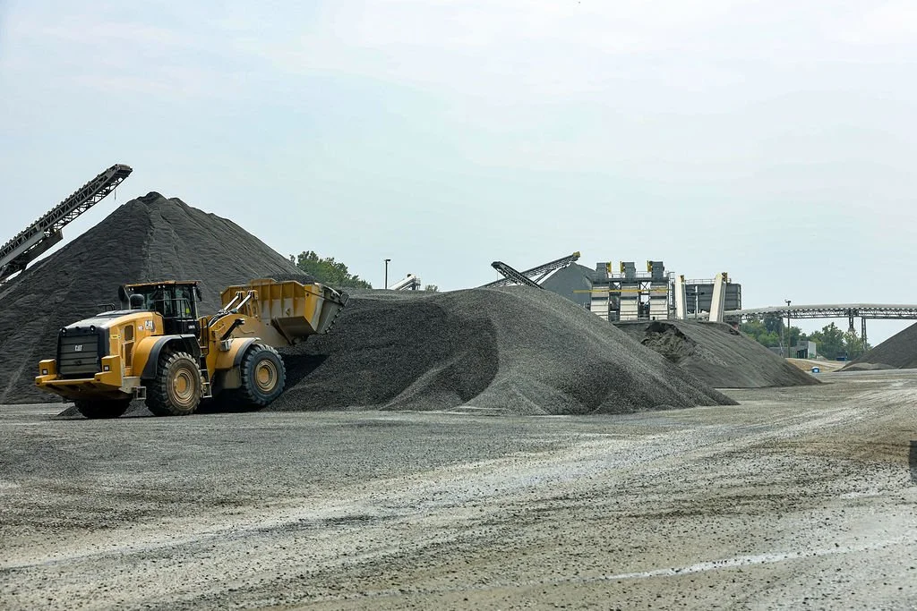 Creekstone construction site with a backhoe loader moving dirt or gravel, large material piles, and industrial structures in the background, highlighting excavation and site-work operations.