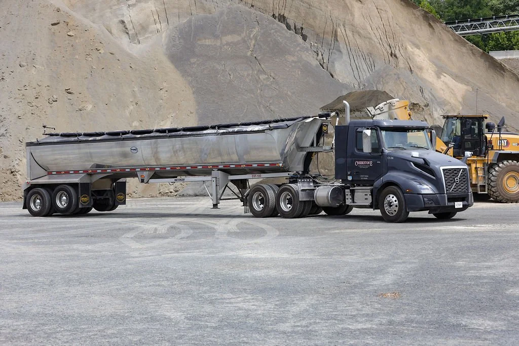 A large black semi-truck with a metallic tanker trailer parked on a gravel lot with a construction excavator in the background.