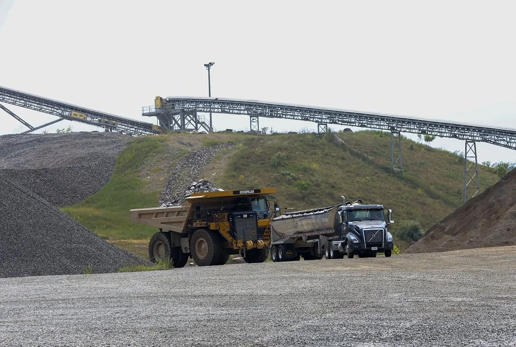 “Creekstone gravel quarry site with heavy machinery, including a dump truck and semi-truck, and a conveyor system on a hill in the background, highlighting aggregate production and material hauling operations.