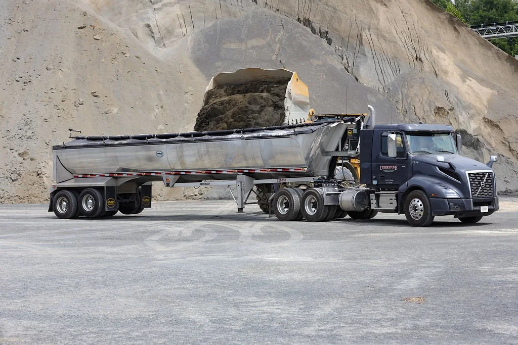 Creekstone dump truck unloading dirt into a semi-truck trailer at an outdoor quarry-style construction site with a dirt hillside in the background, highlighting heavy hauling and material handling operations.