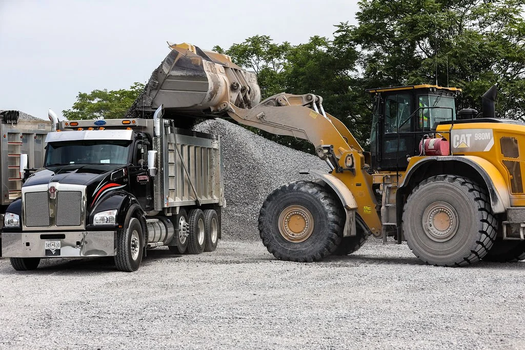 Creekstone yellow Caterpillar front loader loading gravel into a black semi-truck with a silver trailer at a construction site, highlighting heavy equipment operation and material hauling services.