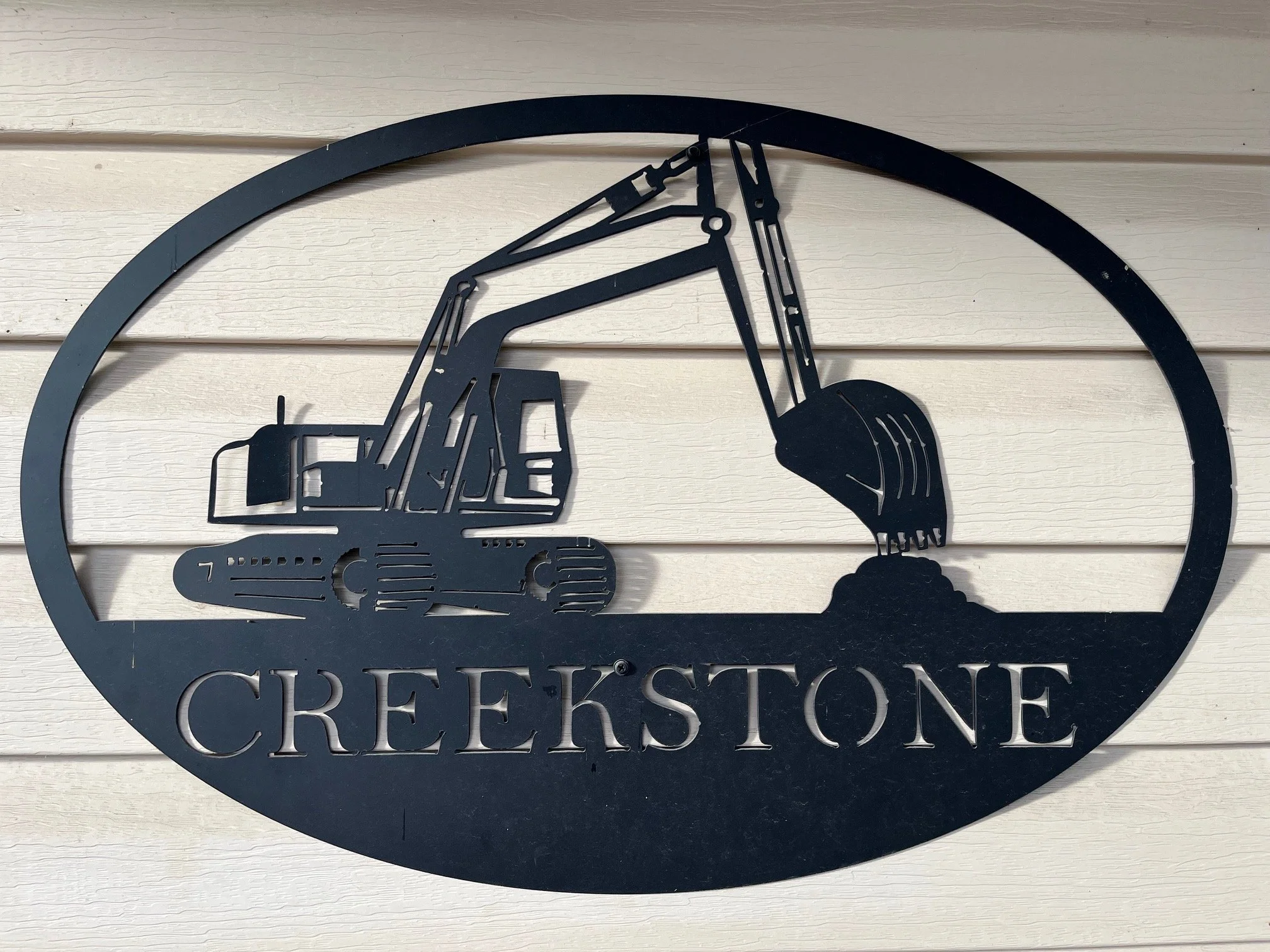 Decorative metal sign of an excavator excavating soil, with the word 'CREEKSTONE' beneath it, mounted on a white wooden wall.