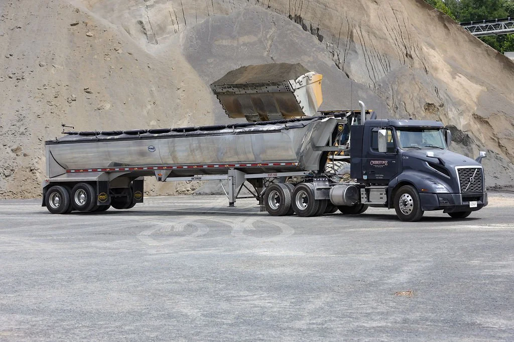 A black semi-truck with a silver trailer is parked on a gravel surface in front of a dirt hill. The trailer is tilted as it unloads a pile of material, possibly gravel or sand, into a conveyor or collection system.