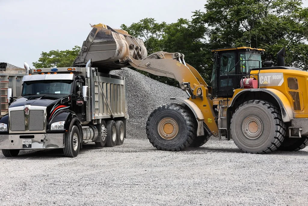 Creekstone yellow CAT construction loader scooping and loading gravel into a black and silver truck at a construction site with a gravel surface and trees in the background, highlighting heavy equipment operation and material hauling services.