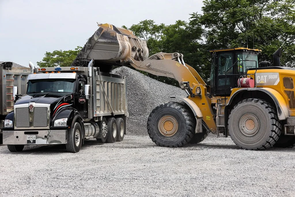 Creekstone yellow Caterpillar front loader transferring gravel into the bed of a black and silver semi-truck, highlighting heavy equipment operation and material hauling services.