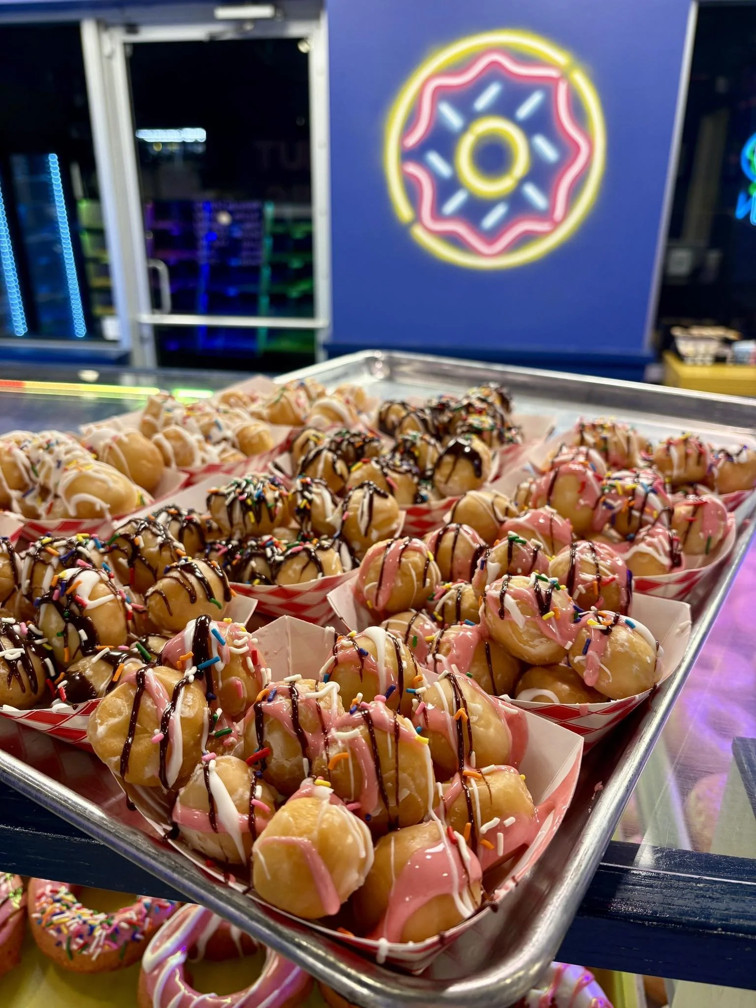 Multiple trays sit on a sheet pan. Each tray has a pile of round donut holes decorated with a variety of icing drizzle and multiple color sprinkles