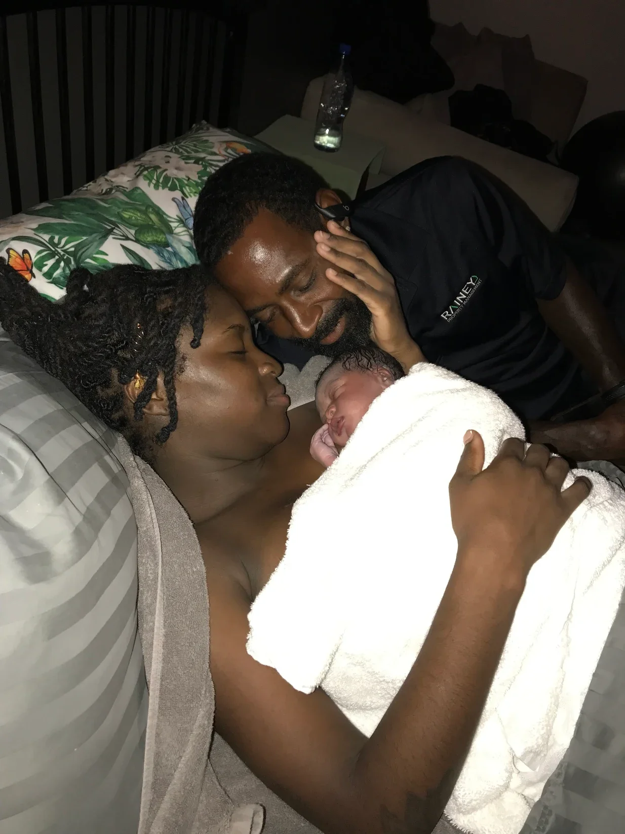 A couple lying in bed holding newborn baby, with an embrace and tender expressions, in a dimly lit room.