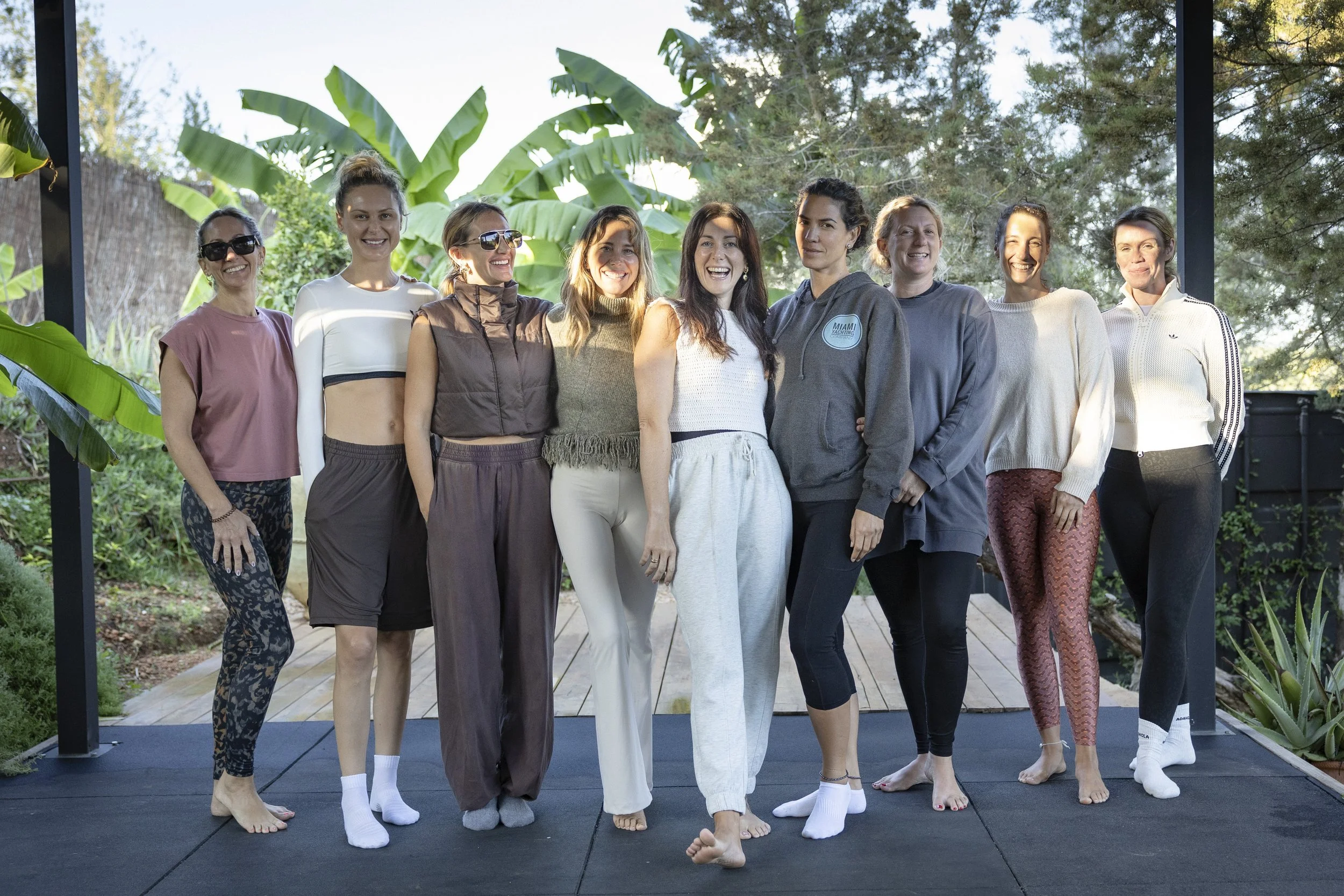 Group of women smiling and standing together on an outdoor stage surrounded by greenery.