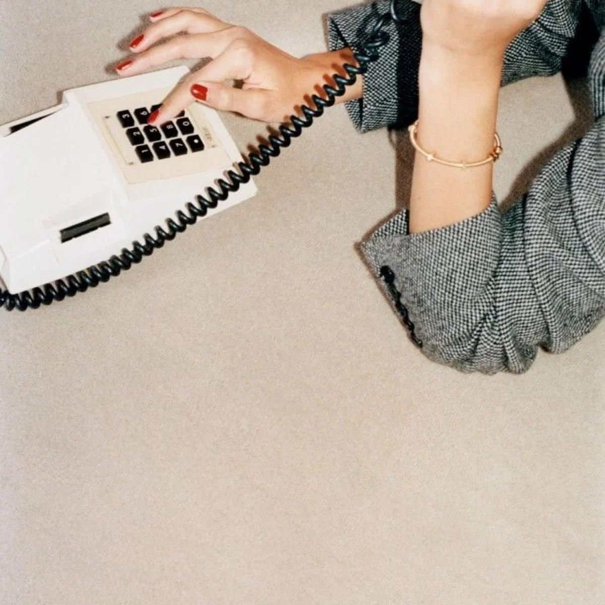 Person with red nail polish and a gold bracelet using a landline phone with a coiled cord on a beige desk.