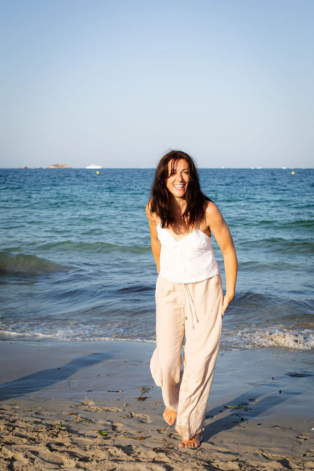 A woman in white and beige clothing smiling and walking on the beach with ocean waves behind her and ships visible on the horizon.