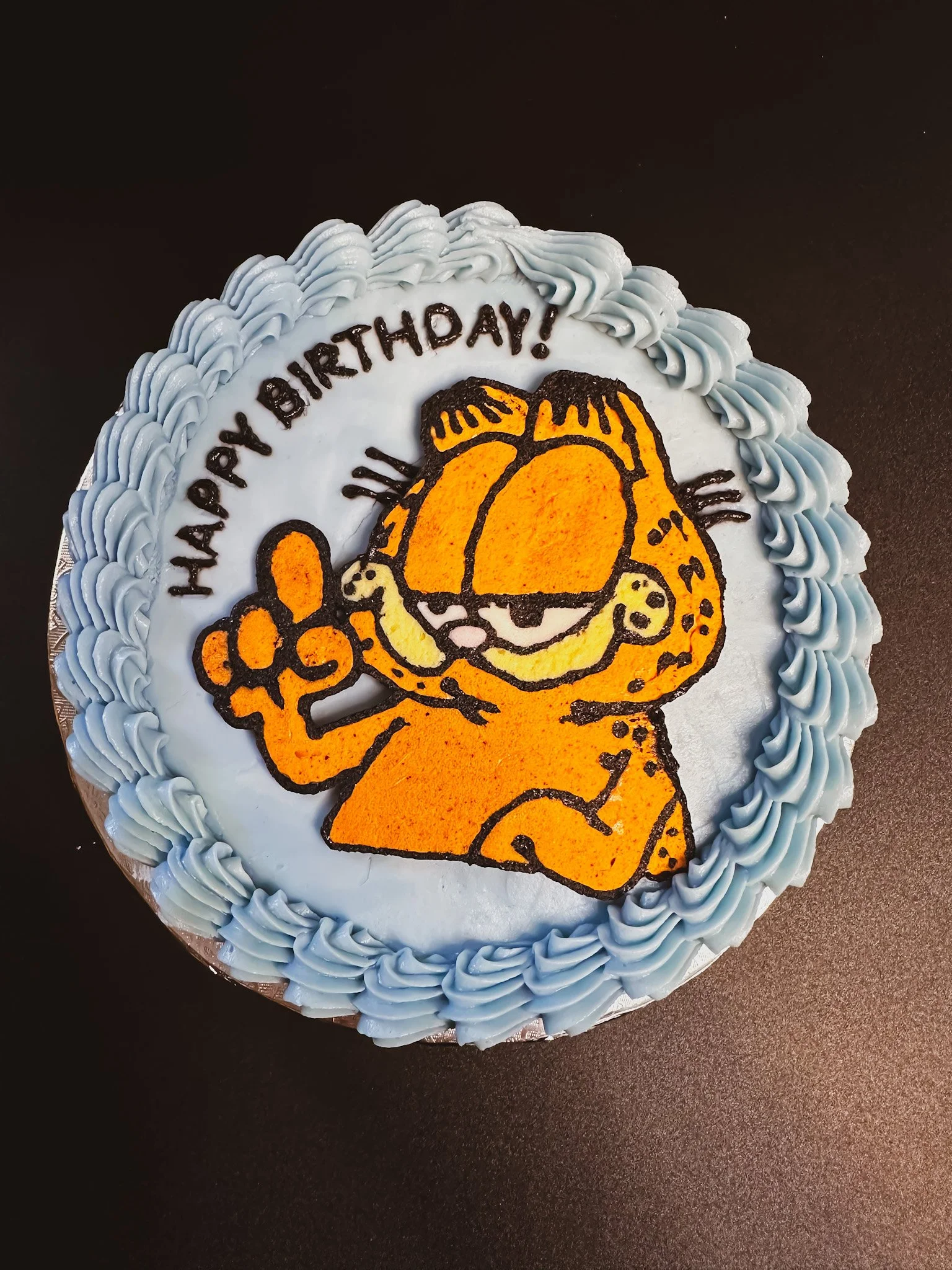 A birthday cake decorated with blue icing and an image of Garfield, the orange cartoon cat, making a peace sign. The cake has the words 'HAPPY BIRTHDAY!' written on it.