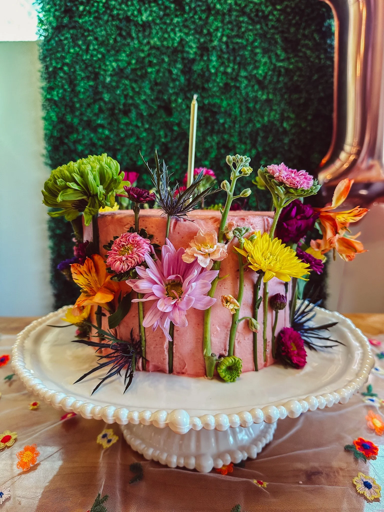 A pink birthday cake decorated with an assortment of colorful flowers on a white cake stand, with a green hedge background and a balloon in the corner.