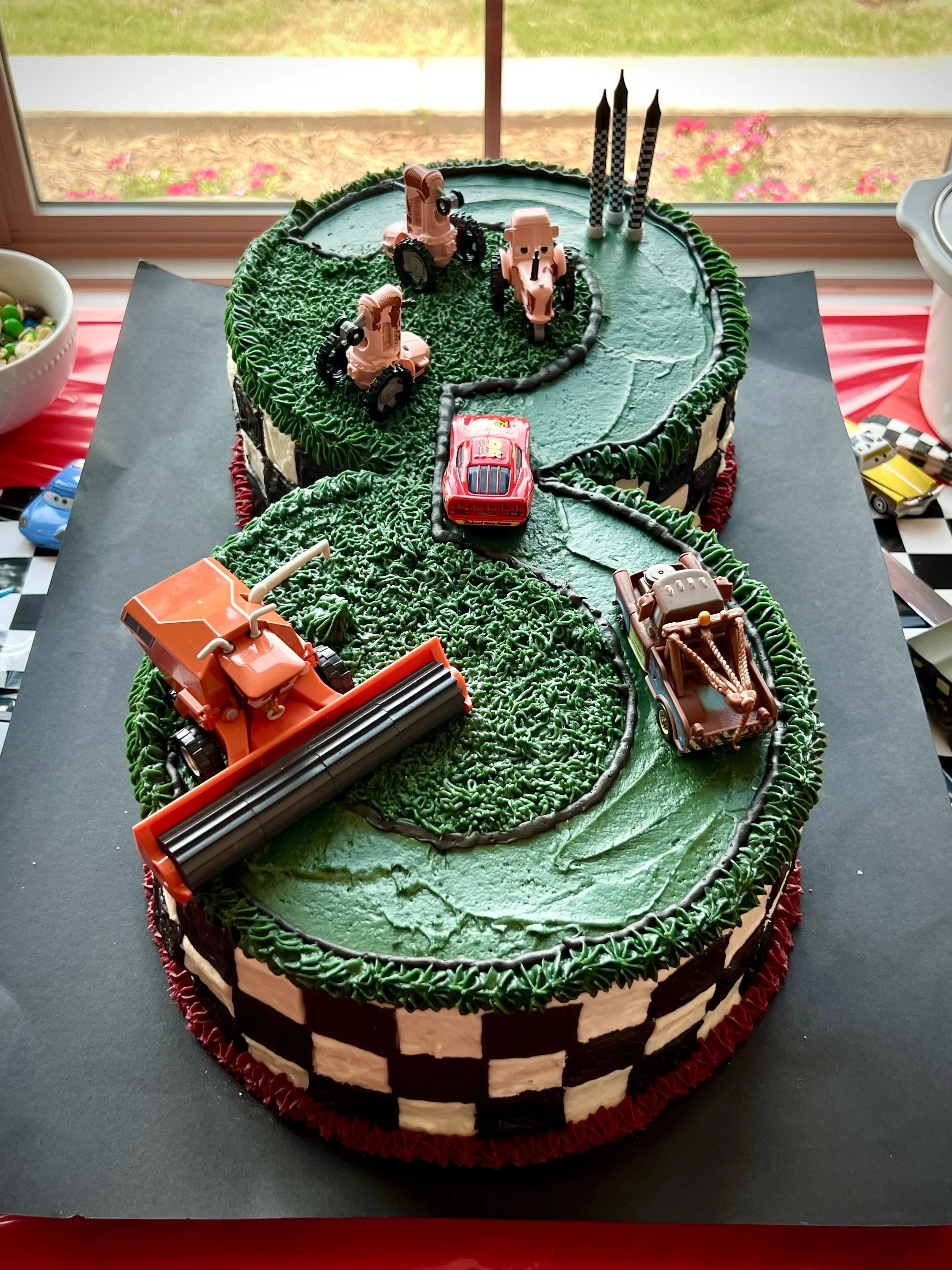Race car-themed birthday cake with green frosting, featuring toy construction and race cars on a checkered pattern border, placed on a black cake board near a window.