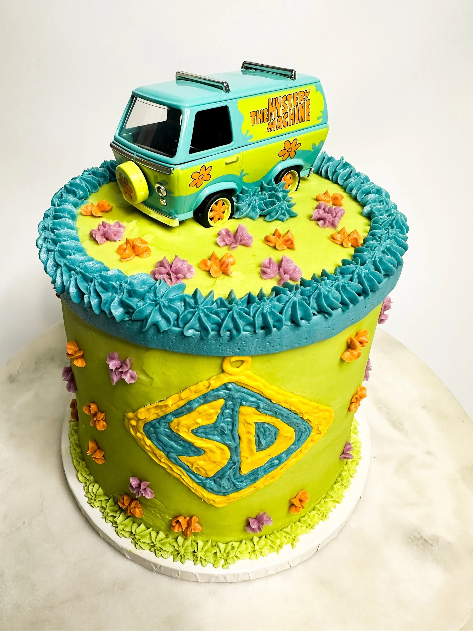 Colorful two-tier cake with a "Mystery Machine" cookie on top and a Superman logo on the side, decorated with purple and orange flowers and blue and green icing.