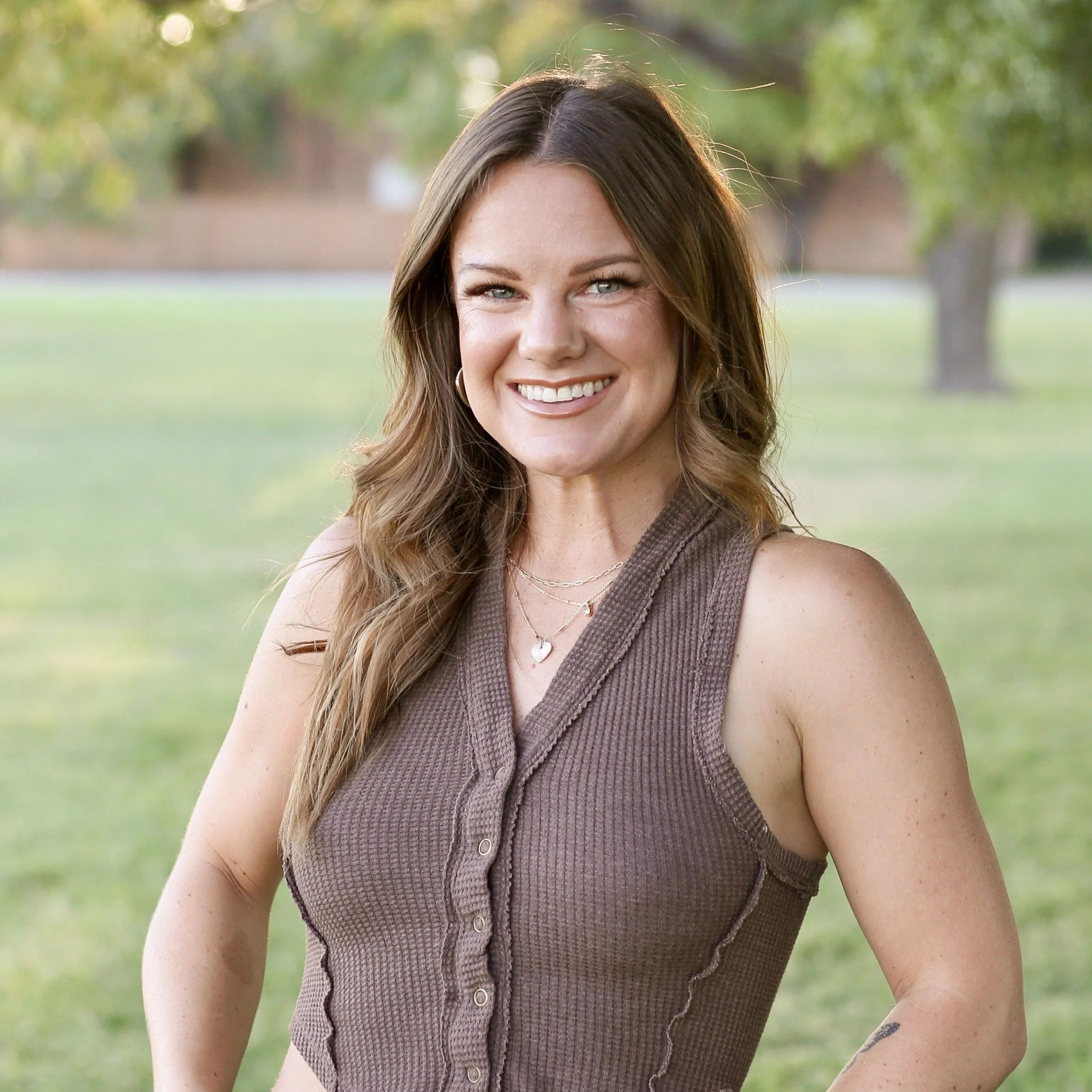 A smiling woman with long wavy brown hair standing outdoors in a park with green grass and trees in the background.