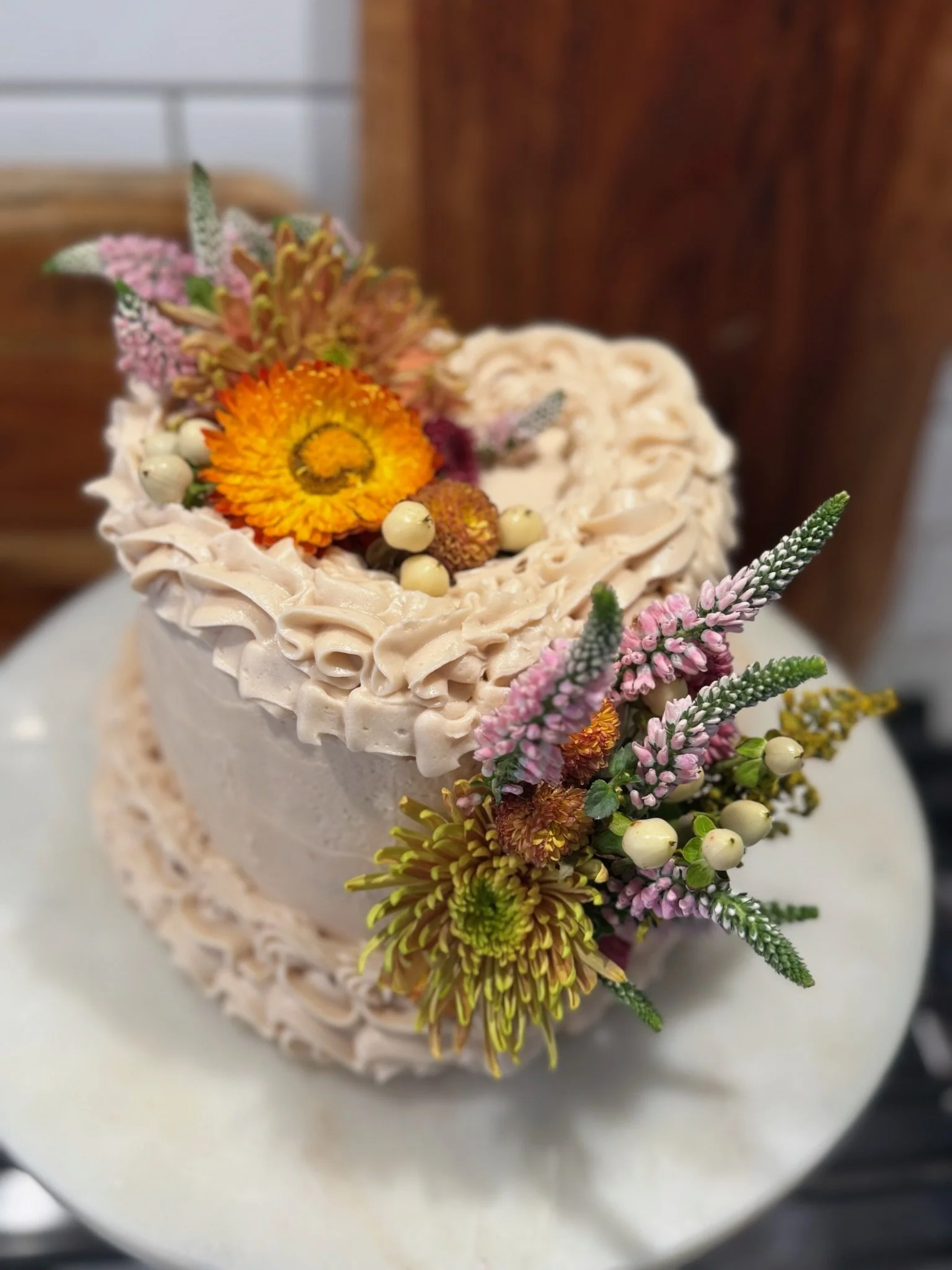 A round, two-tiered cake decorated with pinkish-beige frosting and fresh flowers including orange, yellow, pink, purple, and green blooms, with the top tier featuring a wreath of flowers around the center.