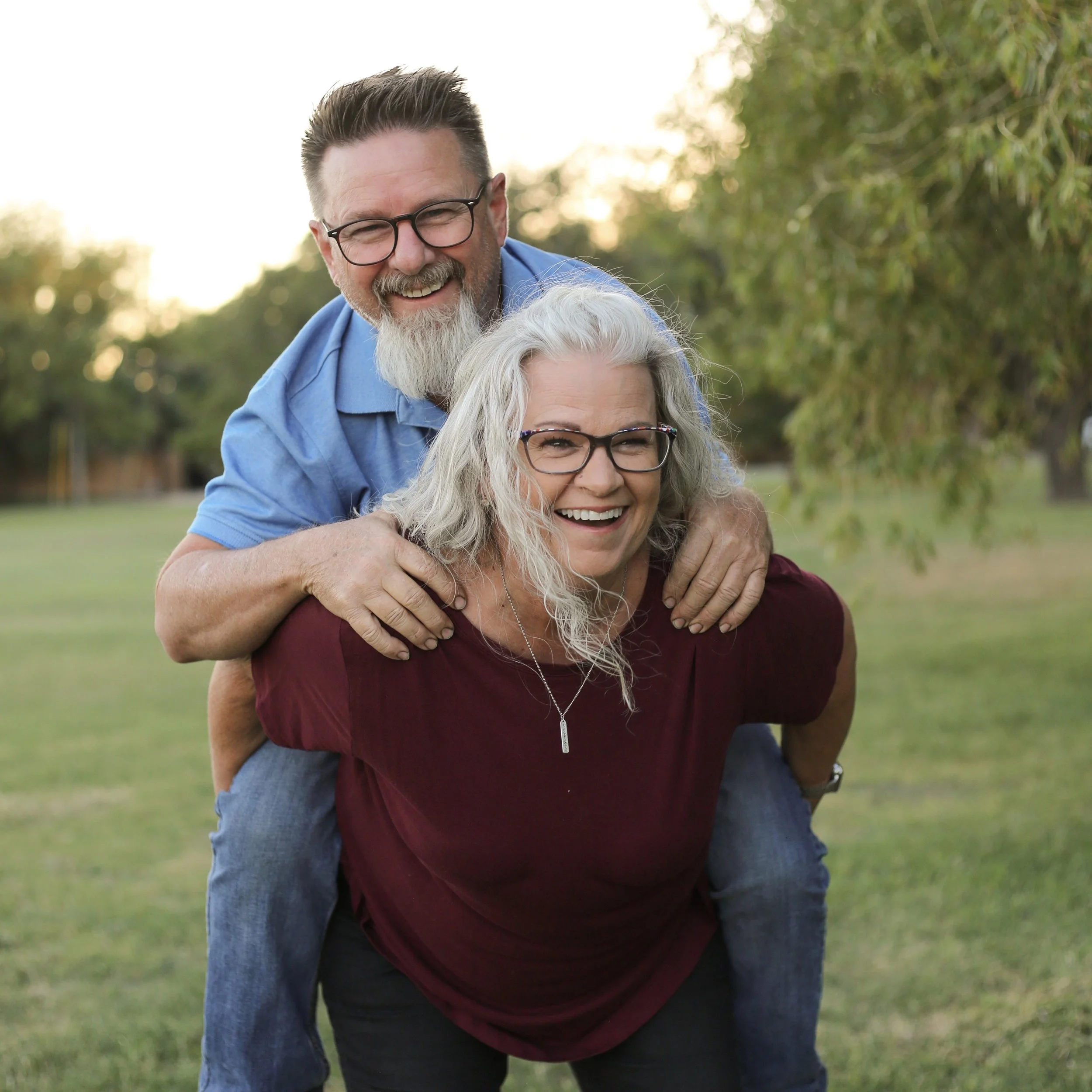 A laughing couple, with the woman giving a piggyback ride to the man in a park with trees and grass.