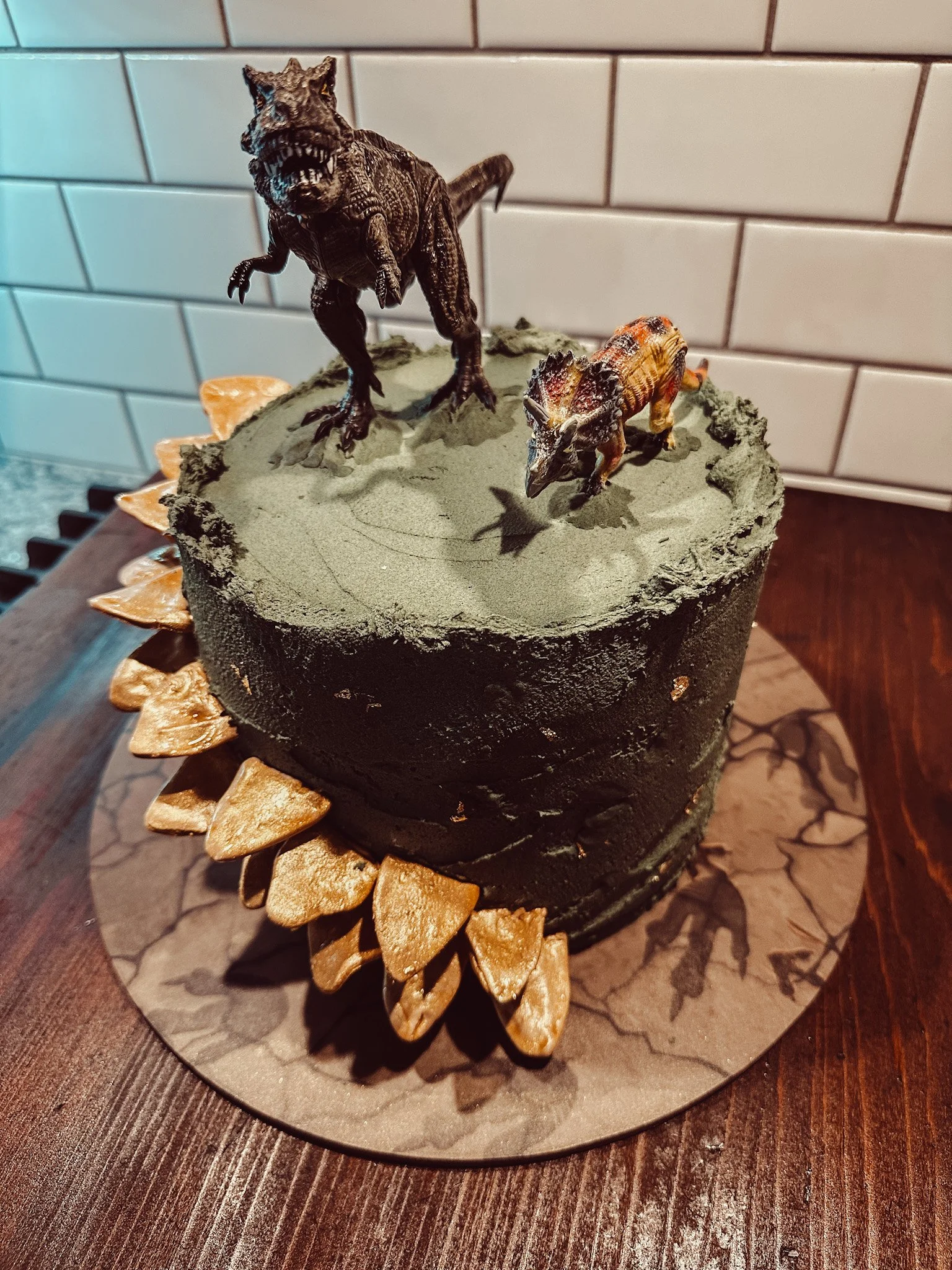 A cake decorated to look like a dinosaur scene. It features a miniature T-Rex, a smaller dinosaur, both on a green topping that resembles mud or grass. The cake has a black exterior with gold-colored edible spikes around the sides, and the scene is s
