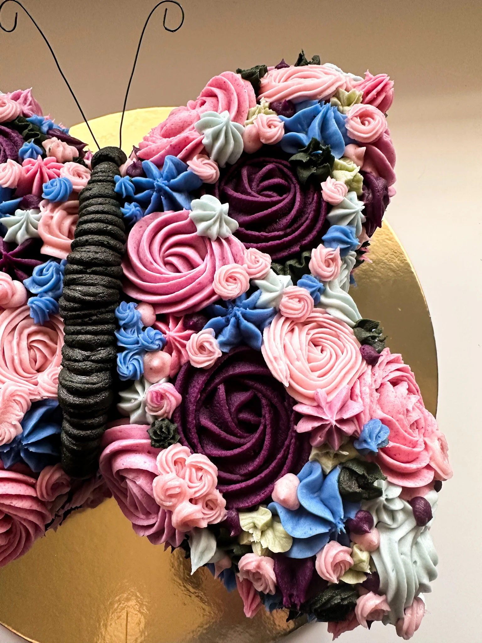 A cake decorated with colorful buttercream flowers forming the shape of a butterfly, with a black licorice antennae detail, on a gold-colored cake board.
