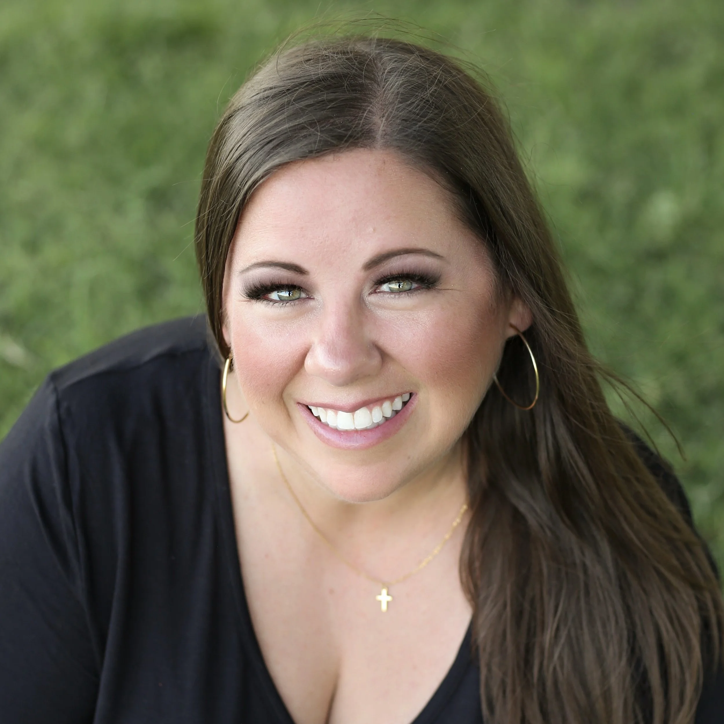A smiling woman with long brown hair, wearing hoop earrings, a cross necklace, and a black top, outdoors with a blurred green background.