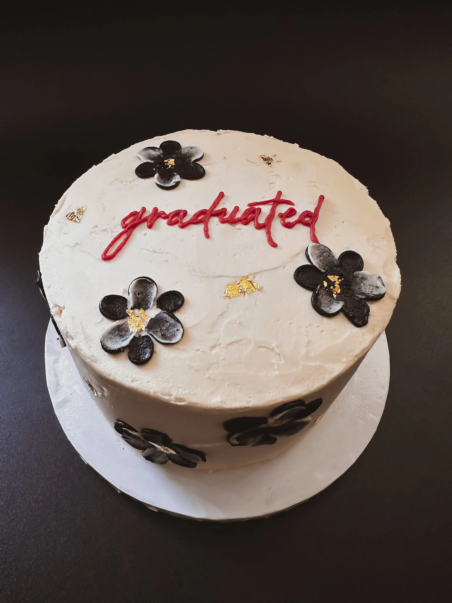 A round white cake decorated with black flowers and gold accents, with the word 'graduated' written in red icing on top.