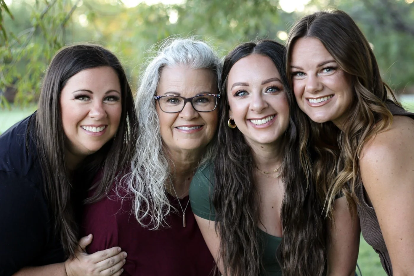 Four women smiling closely together outdoors in a park.