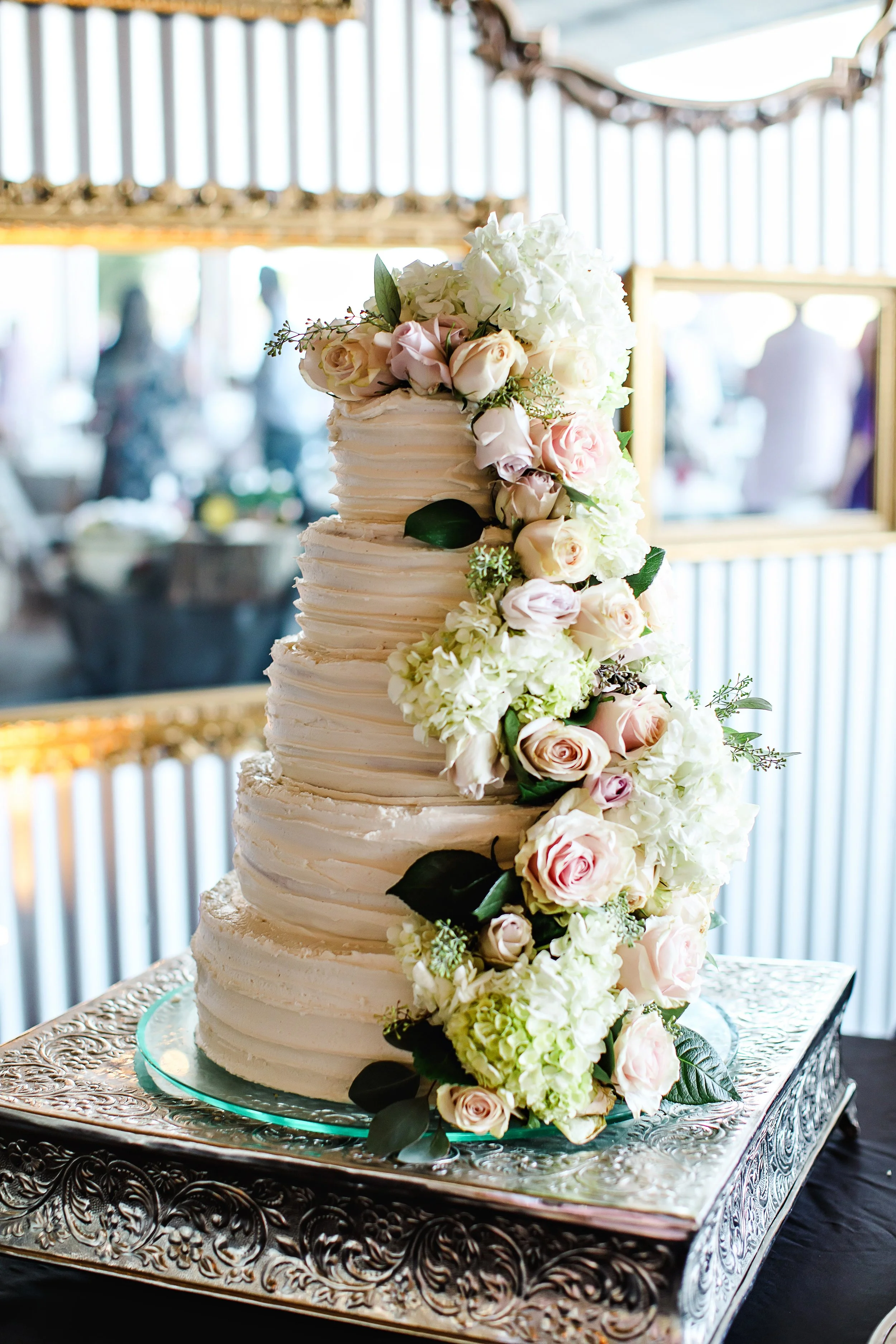 A tall, four-tier wedding cake decorated with white buttercream frosting and cascading pink and white roses with green leaves, placed on an ornate silver cake stand at a wedding reception.