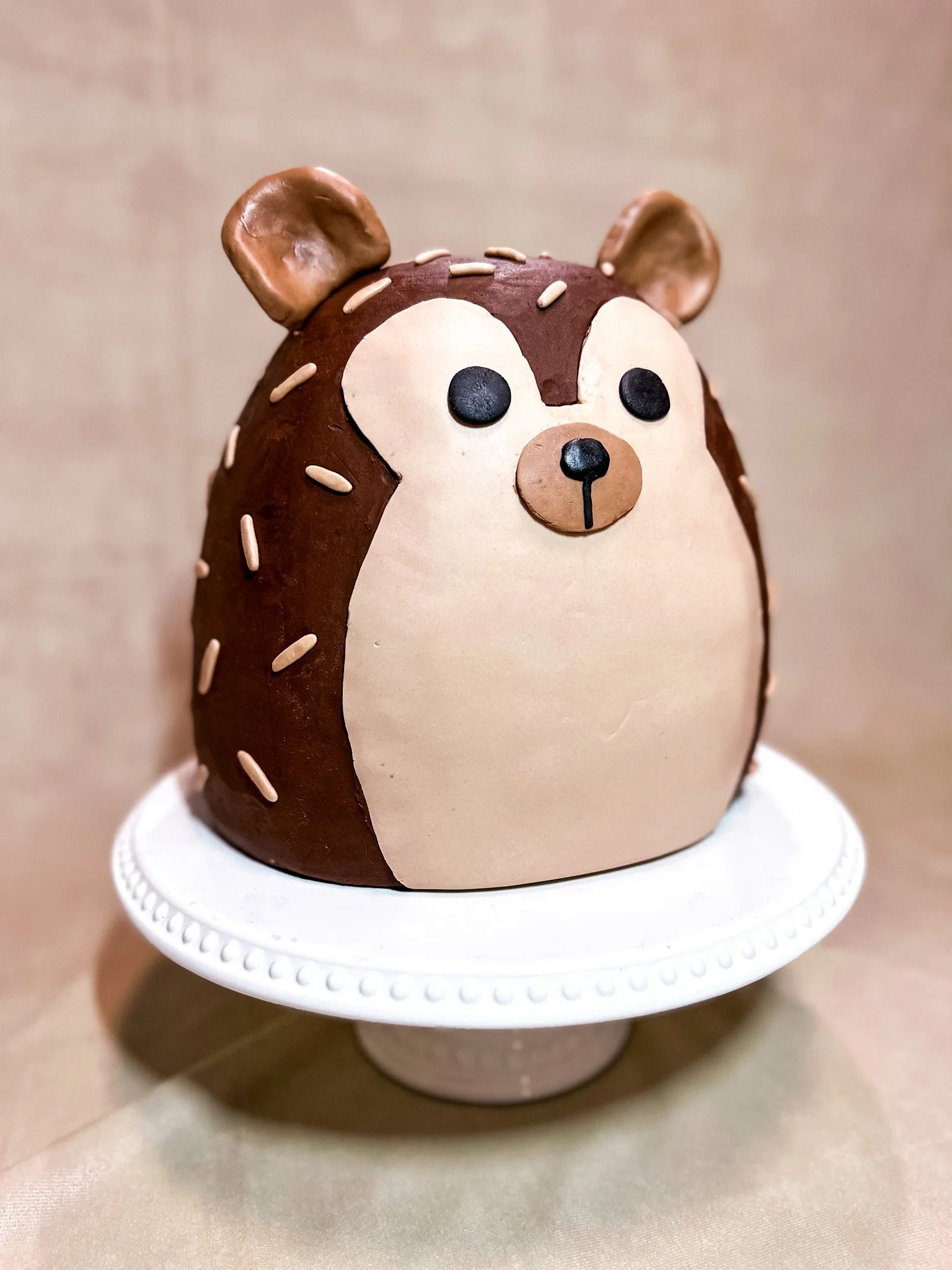 A cake designed to look like a cute hedgehog with chocolate and beige frosting, placed on a white cake stand.