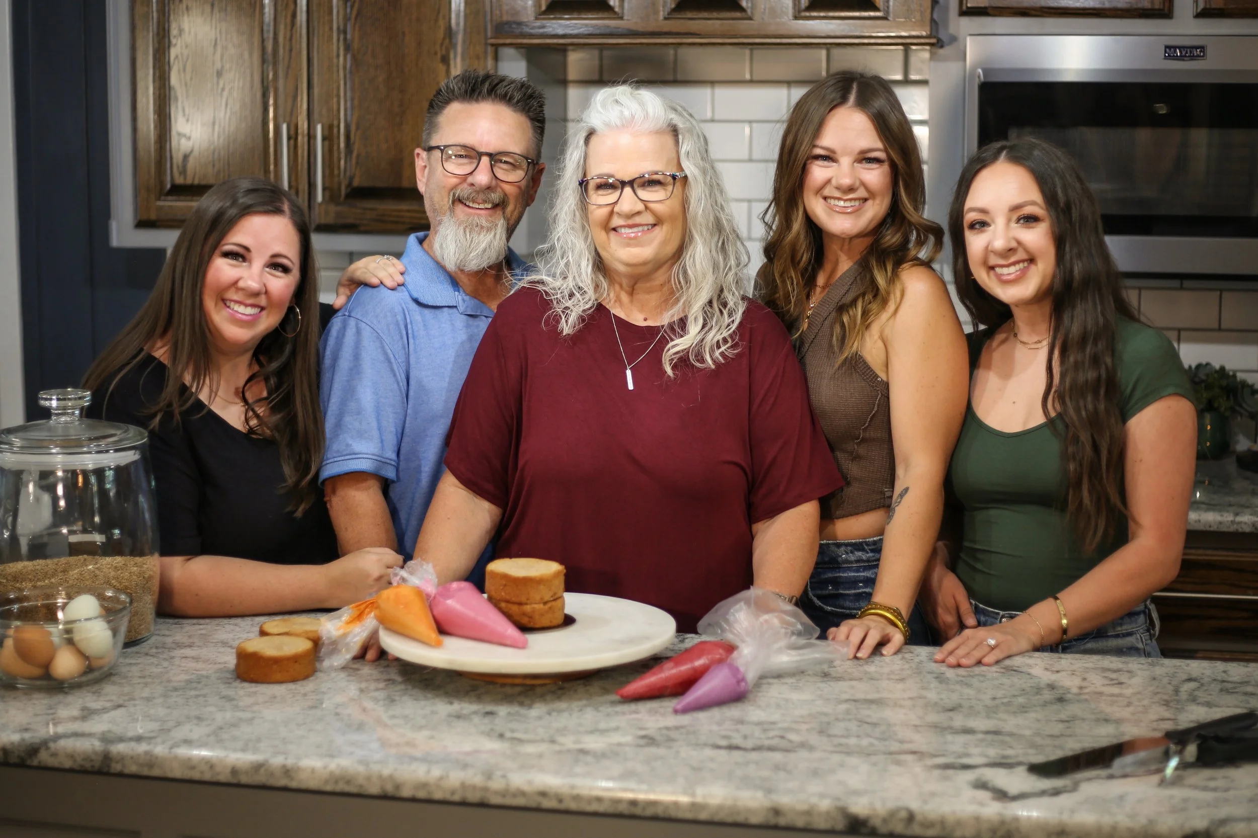 Six smiling people, five women and one man, gathered around a kitchen island with colorful icing bags and baked goods, in a cozy kitchen setting.