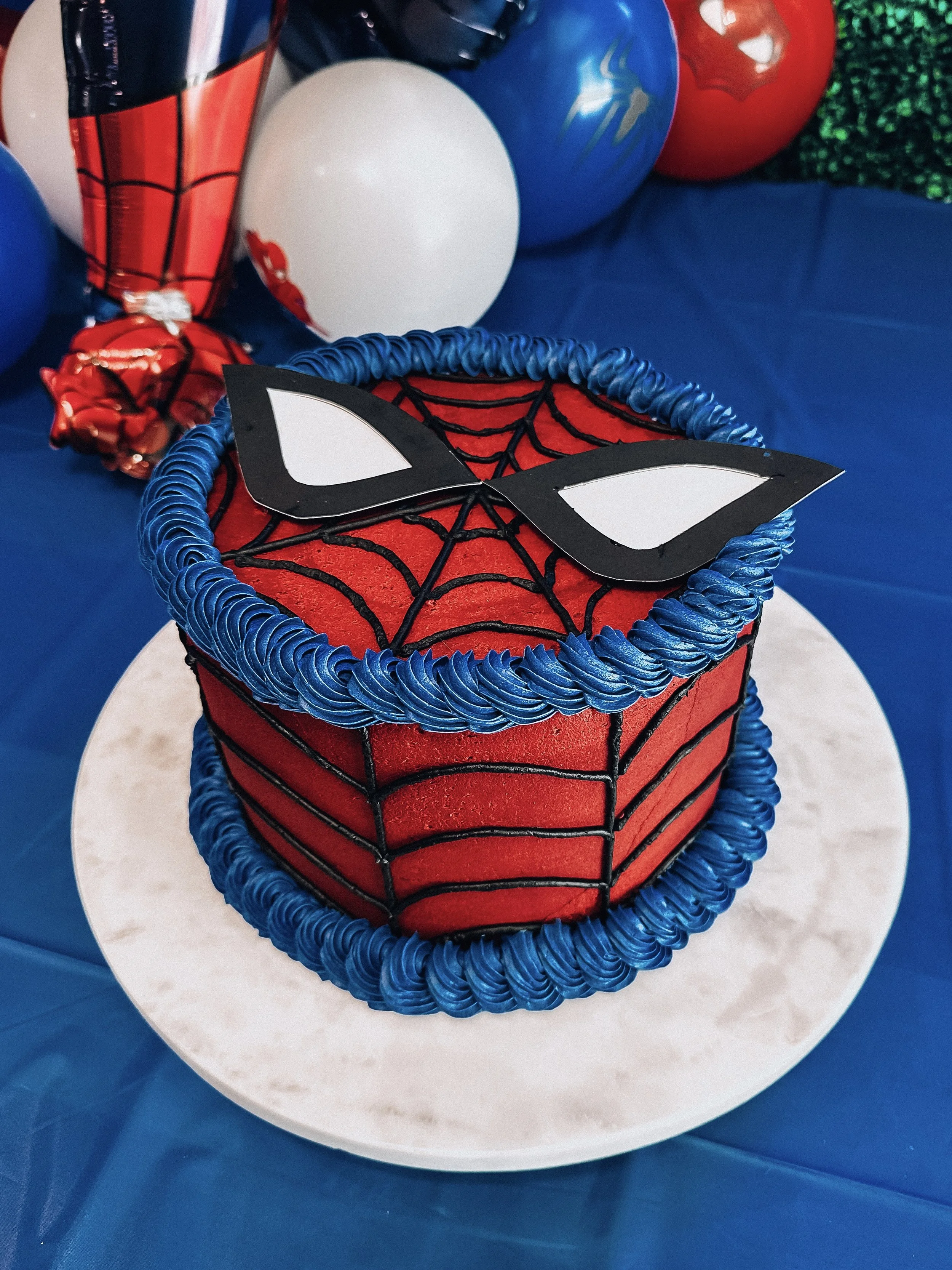 Spider-Man themed birthday cake with red frosting, black web detailing, and Spider-Man mask on top, decorated with blue icing border. Balloons in the background with blue, white, red, and green colors.