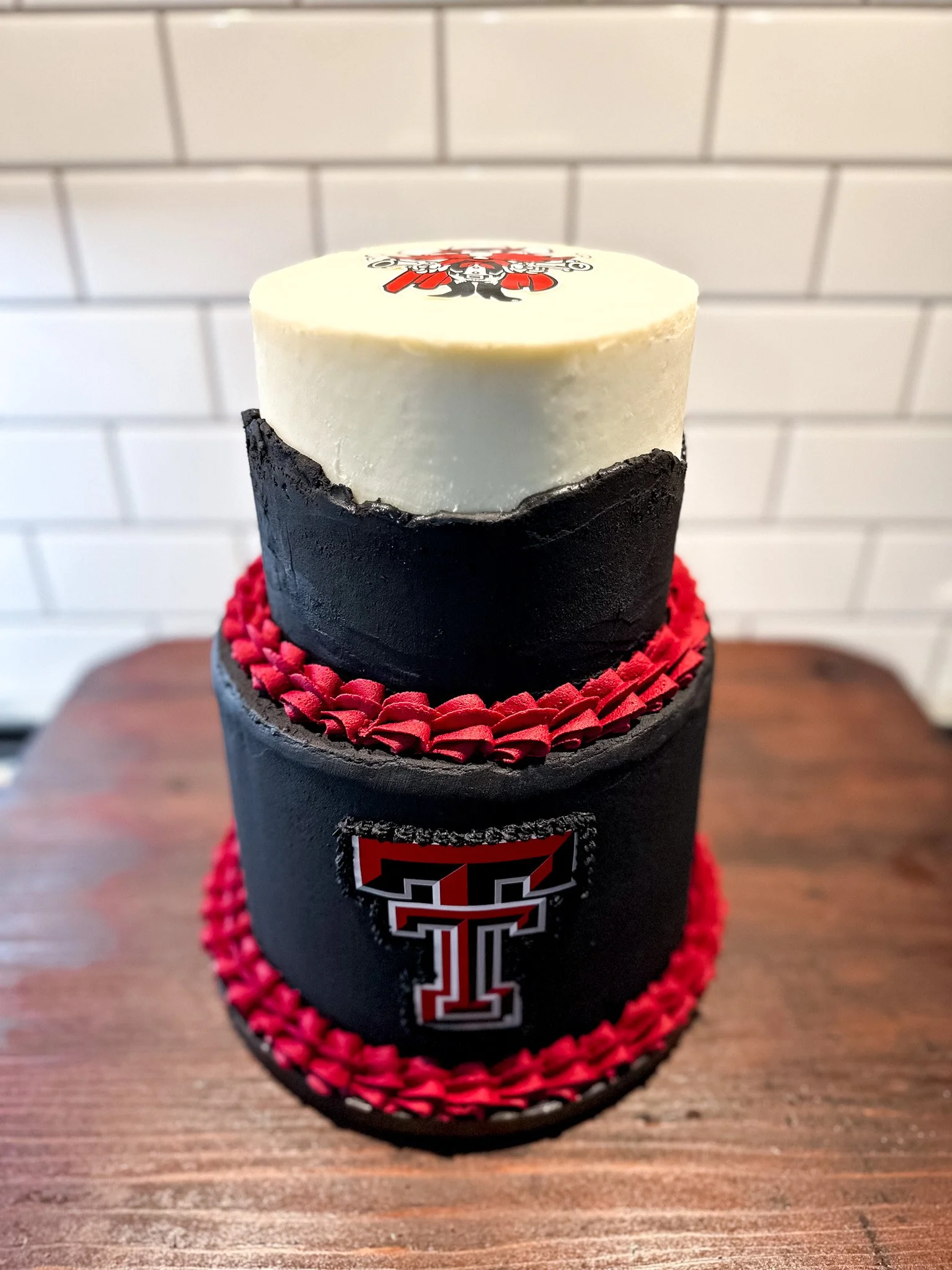 A three-tiered cake decorated with Texas Tech University colors of black, red, and white. The cake features the Texas Tech logo on the middle tier and is topped with a white fondant disc with a Texas Tech emblem. The cake has red and black piping aro