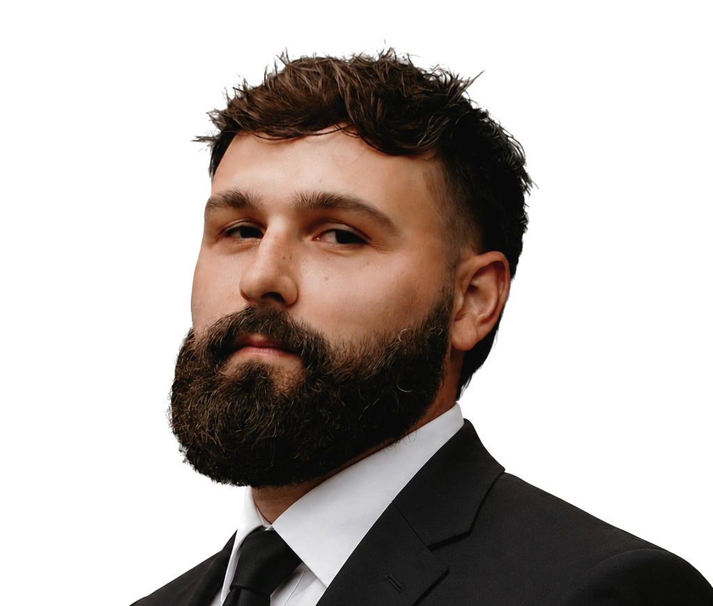 A man with a full beard and styled brown hair, wearing a plaid suit jacket and white dress shirt, looking at the camera with a confident expression.