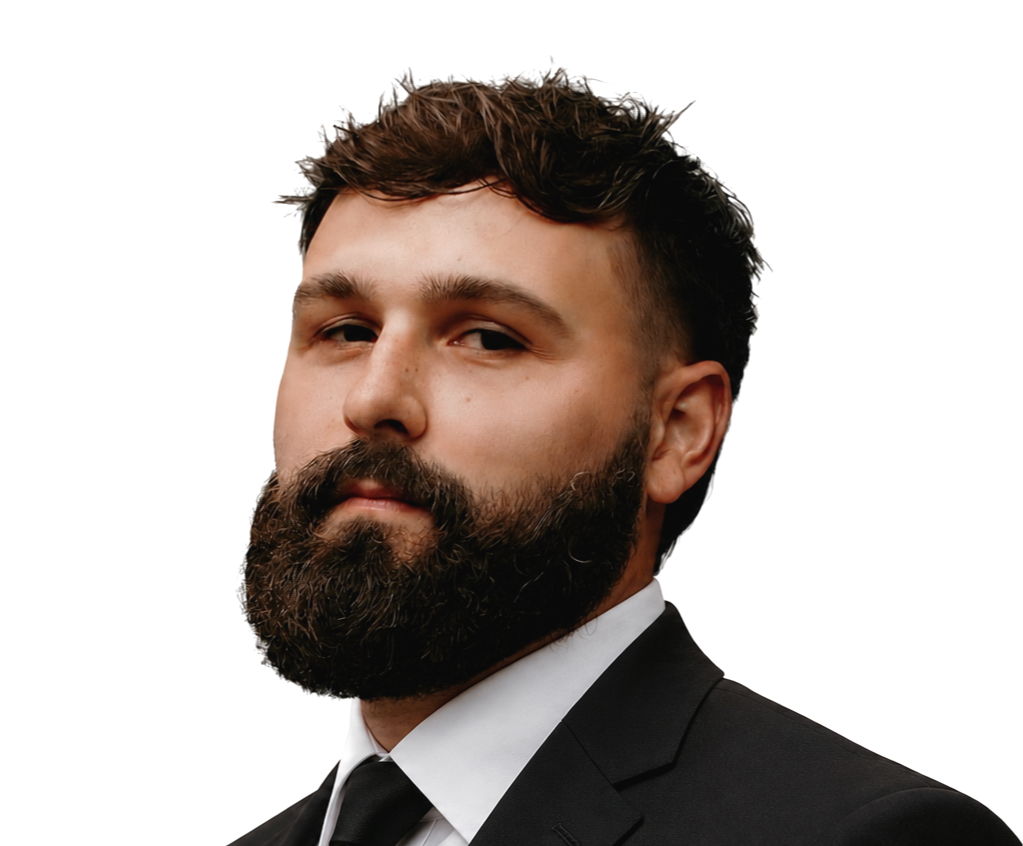 A man with a full beard and styled brown hair, wearing a plaid suit jacket and white dress shirt, looking at the camera with a confident expression.